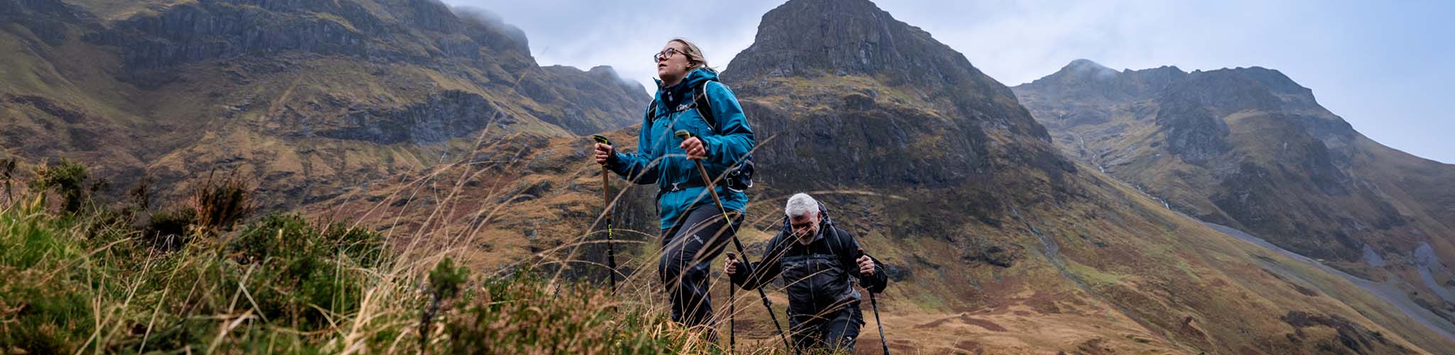 A woman at the front and a man behind her are walking up a hill, wearing jackets and rucksacks and using trekking poles