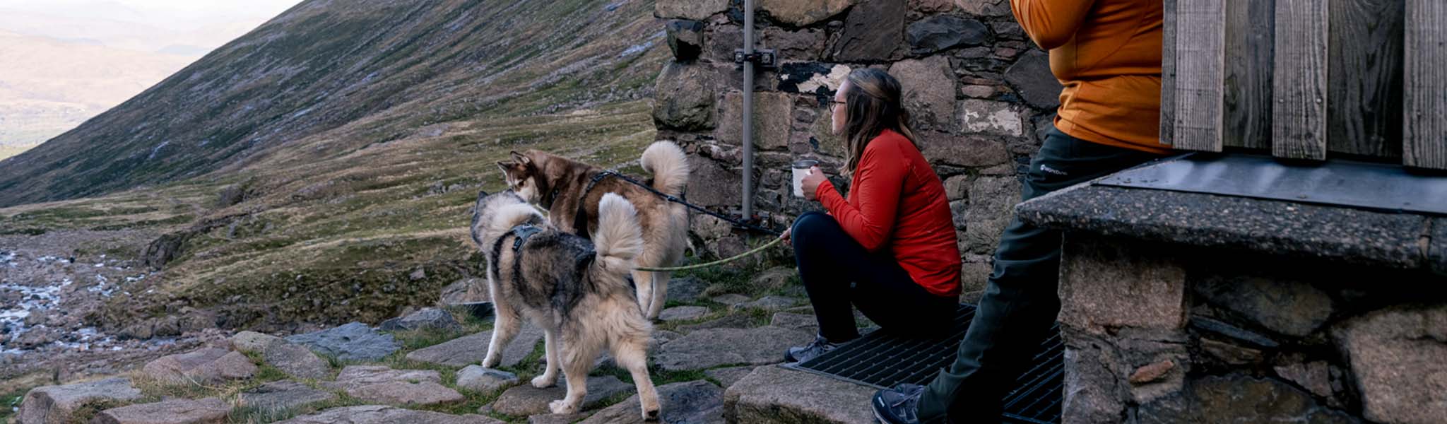 Two people and dogs by a wooden hut are enjoying a cup of tea and outdoors.
